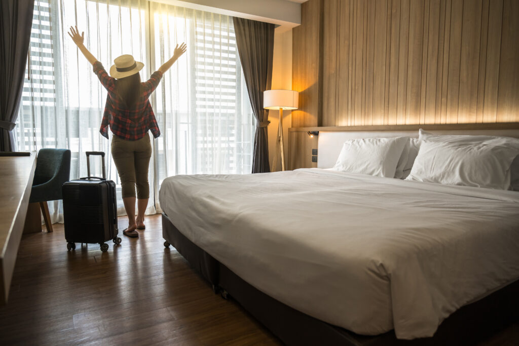 Foto de uma mulher num quarto de hotel com os braços abertos e com sua mala ao lado